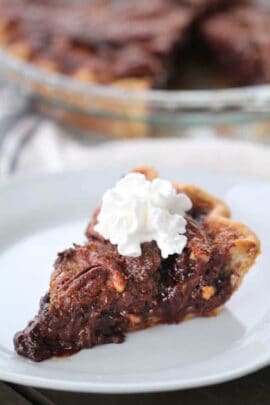 Slice of chocolate pecan pie topped with whipped cream on a white plate.