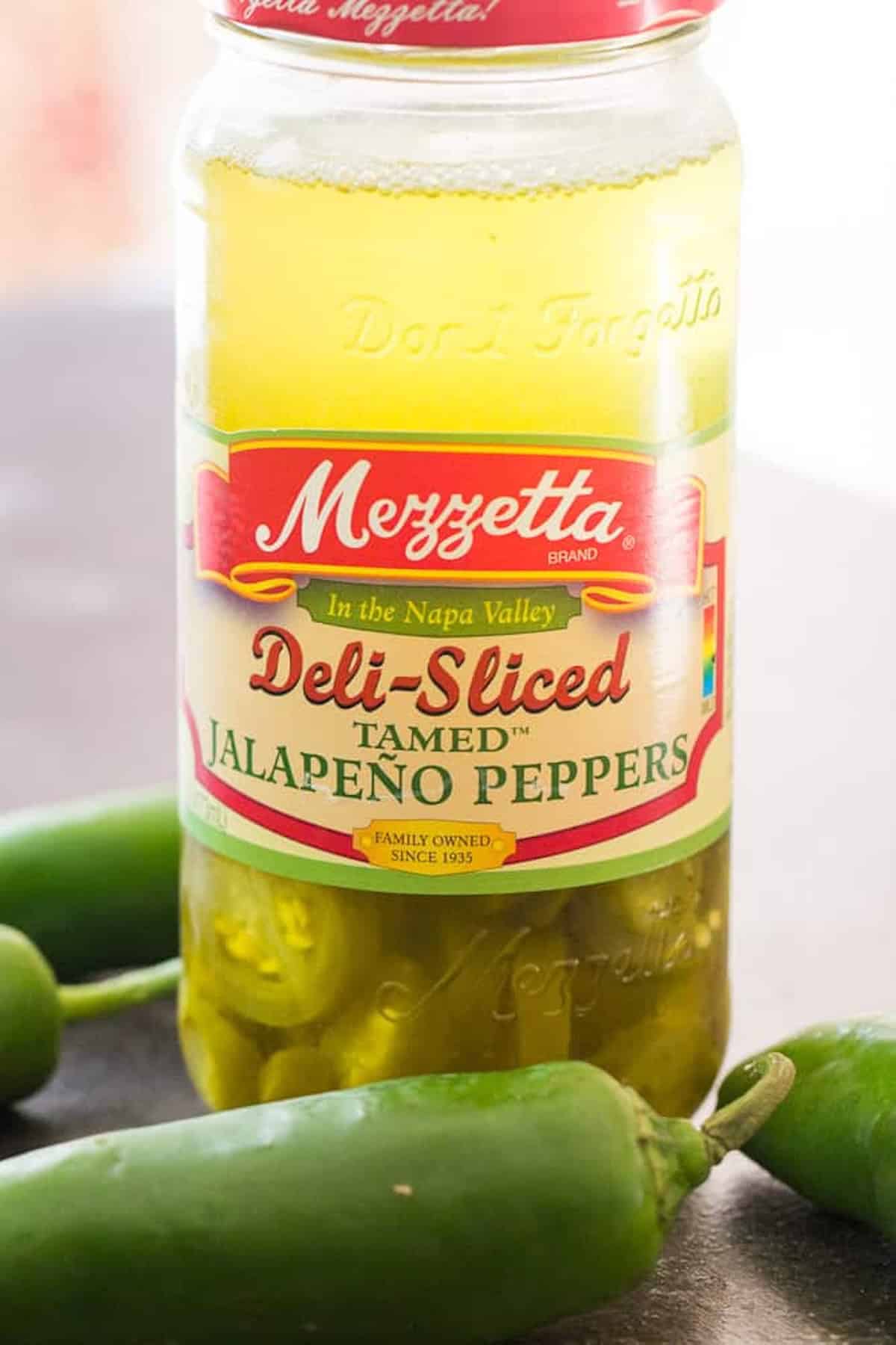 A jar of Mezzetta deli-sliced tamed jalapeño peppers next to fresh jalapeños on a table.