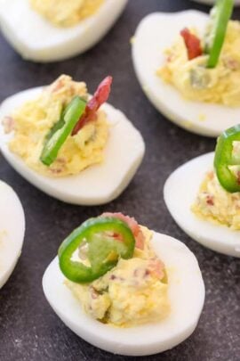 Deviled eggs topped with sliced jalapeños and bacon pieces on a dark surface.