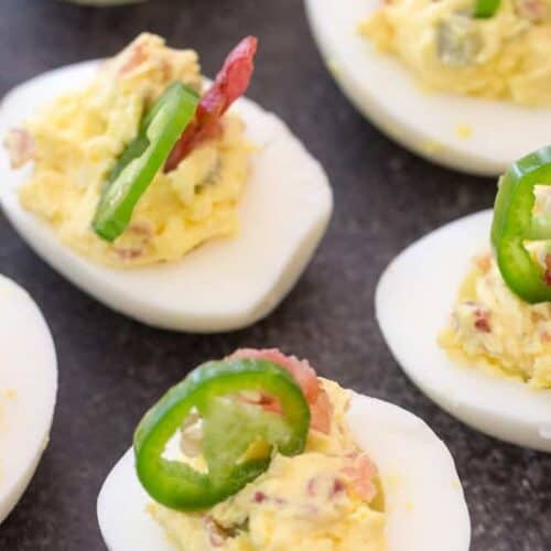 Deviled eggs topped with sliced jalapeños and bacon pieces on a dark surface.