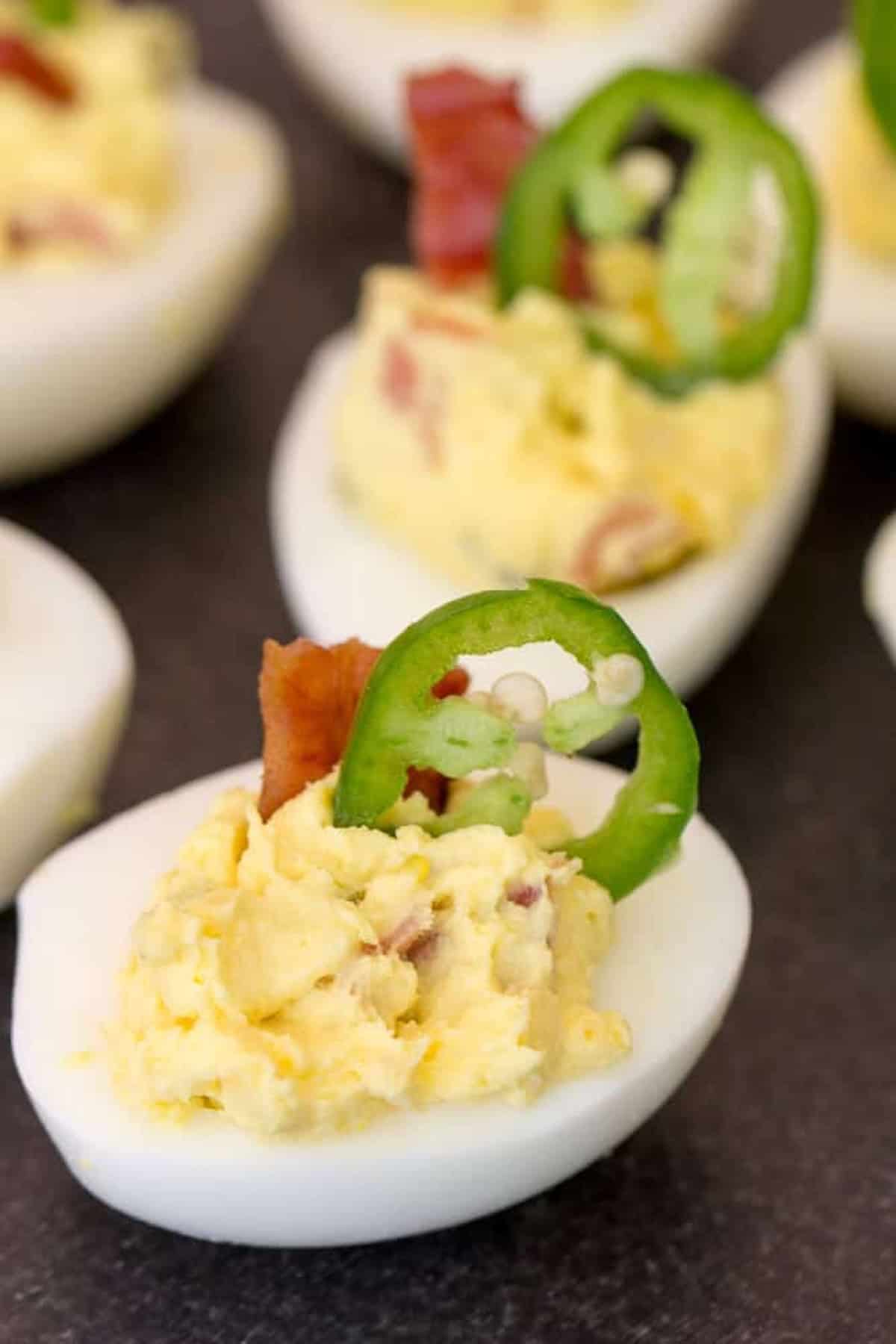 Deviled eggs topped with bacon bits and a slice of jalapeño on a dark background.