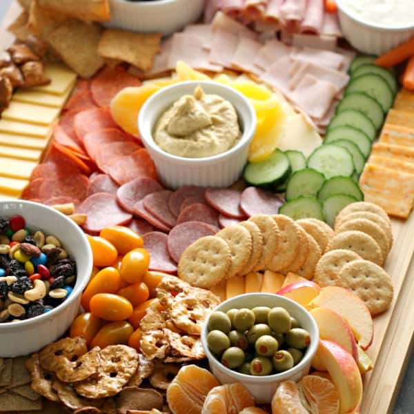 A wooden board filled with assorted snacks, including sliced meats, cheeses, crackers, fresh vegetables, fruits, dips, and nuts arranged in small bowls and sections.