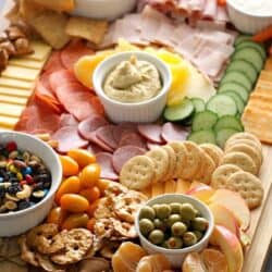 A wooden board filled with assorted snacks, including sliced meats, cheeses, crackers, fresh vegetables, fruits, dips, and nuts arranged in small bowls and sections.