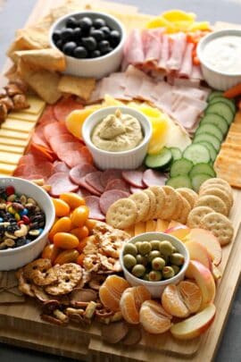 A wooden board filled with assorted snacks, including sliced meats, cheeses, crackers, fresh vegetables, fruits, dips, and nuts arranged in small bowls and sections.