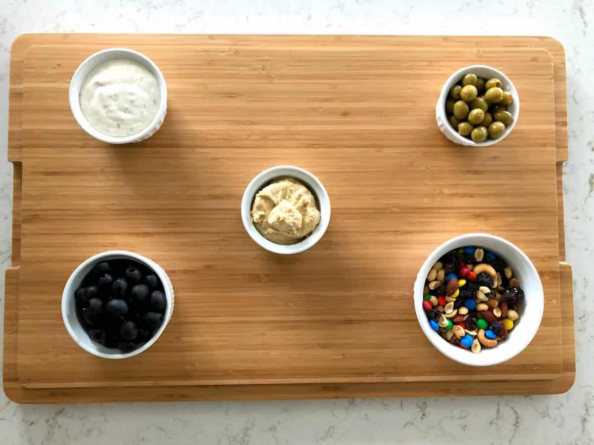 Five small bowls on a wooden board: black olives, mixed nuts with candy, green olives, a central bowl with hummus, and a bowl with dip sauce.