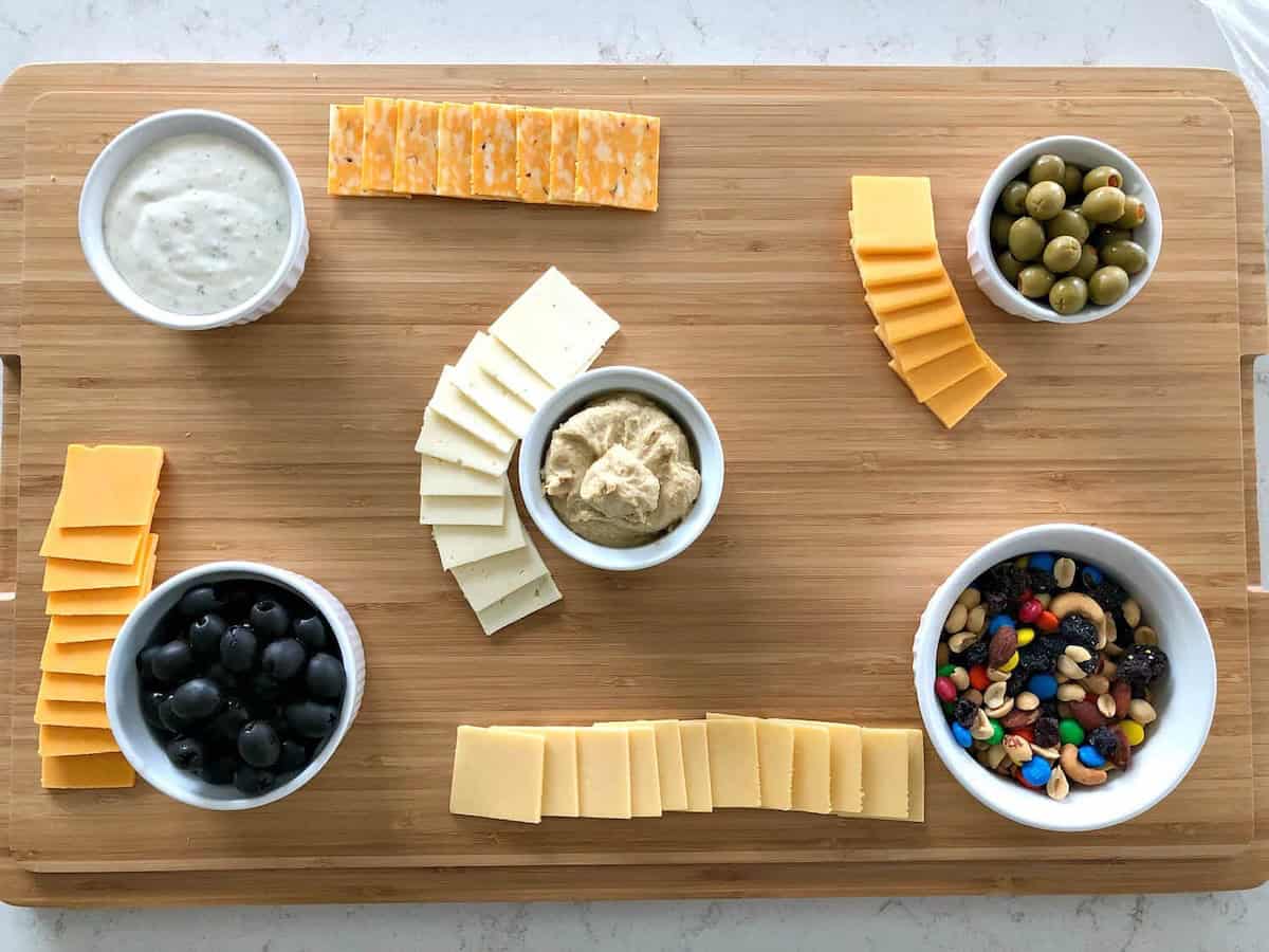 A wooden board with a variety of cheeses, sliced olives, black olives, mixed nuts, and dips in bowls.