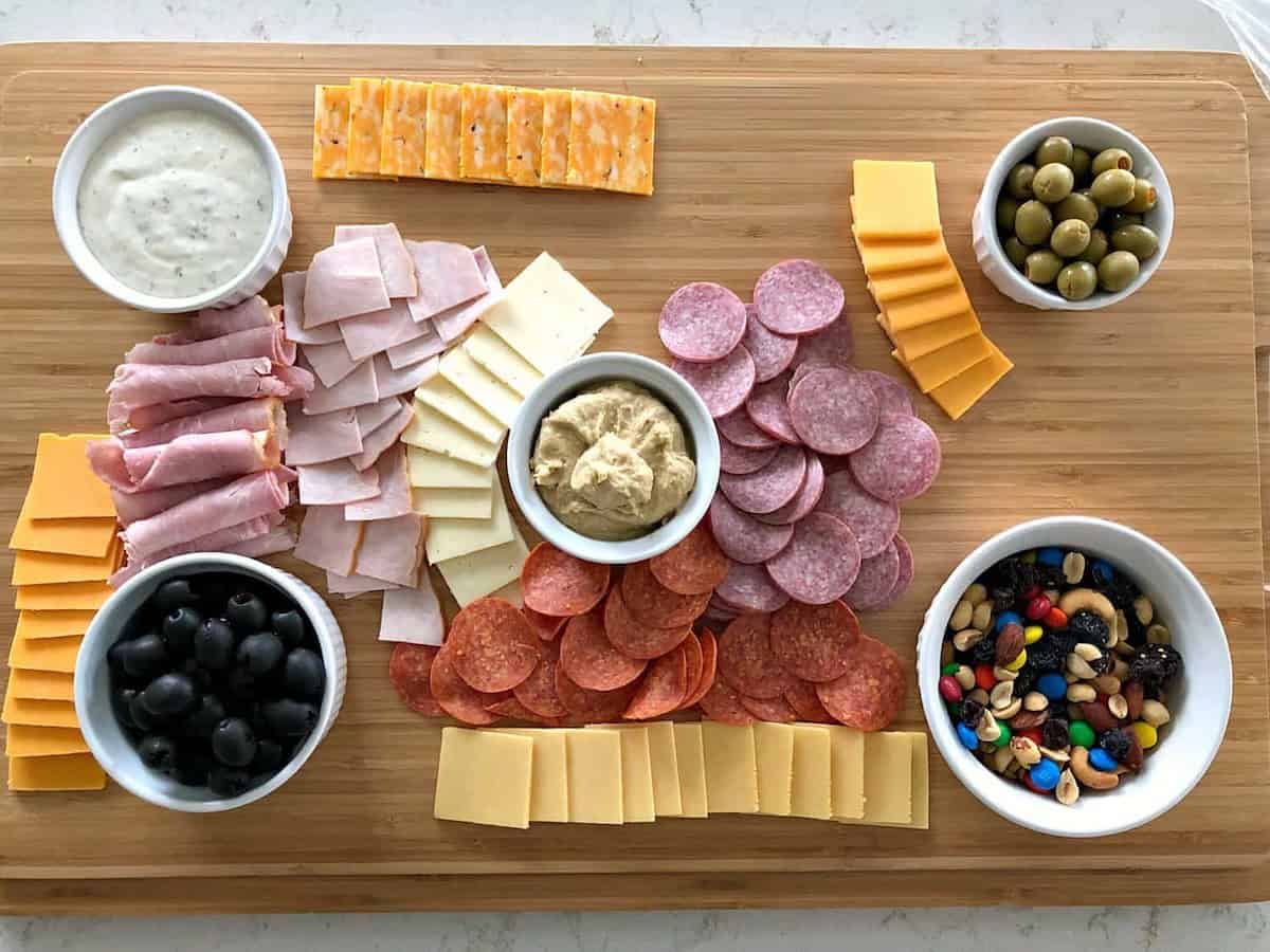 A charcuterie board with sliced meats, various cheeses, olives, nuts, and small bowls of hummus and dips.