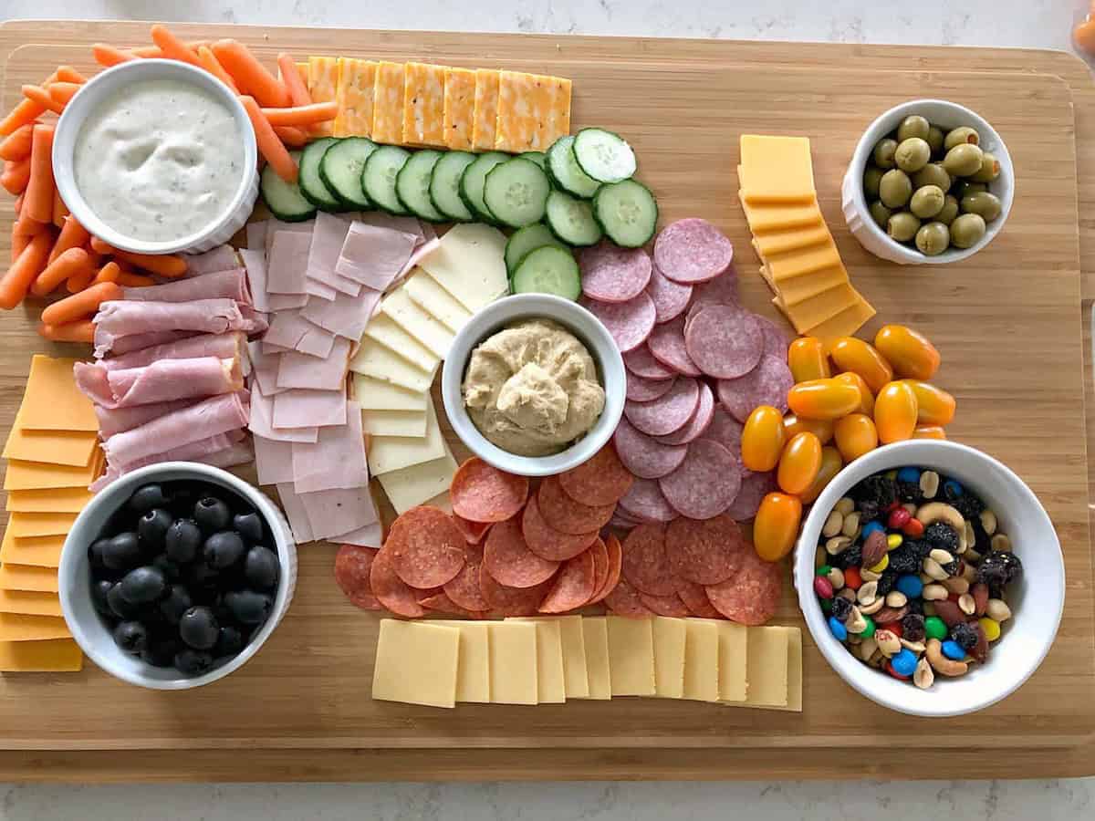 A charcuterie board with assorted meats, cheeses, sliced cucumber, cherry tomatoes, and bowls of olives, nuts, and dips on a wooden surface.