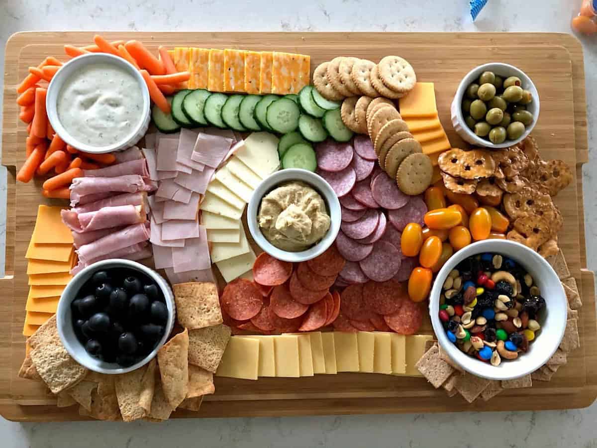 A wooden board with various snacks: sliced meats, cheeses, crackers, olives, sliced cucumber, baby carrots, cherry tomatoes, dips, and a bowl of mixed nuts and candies.