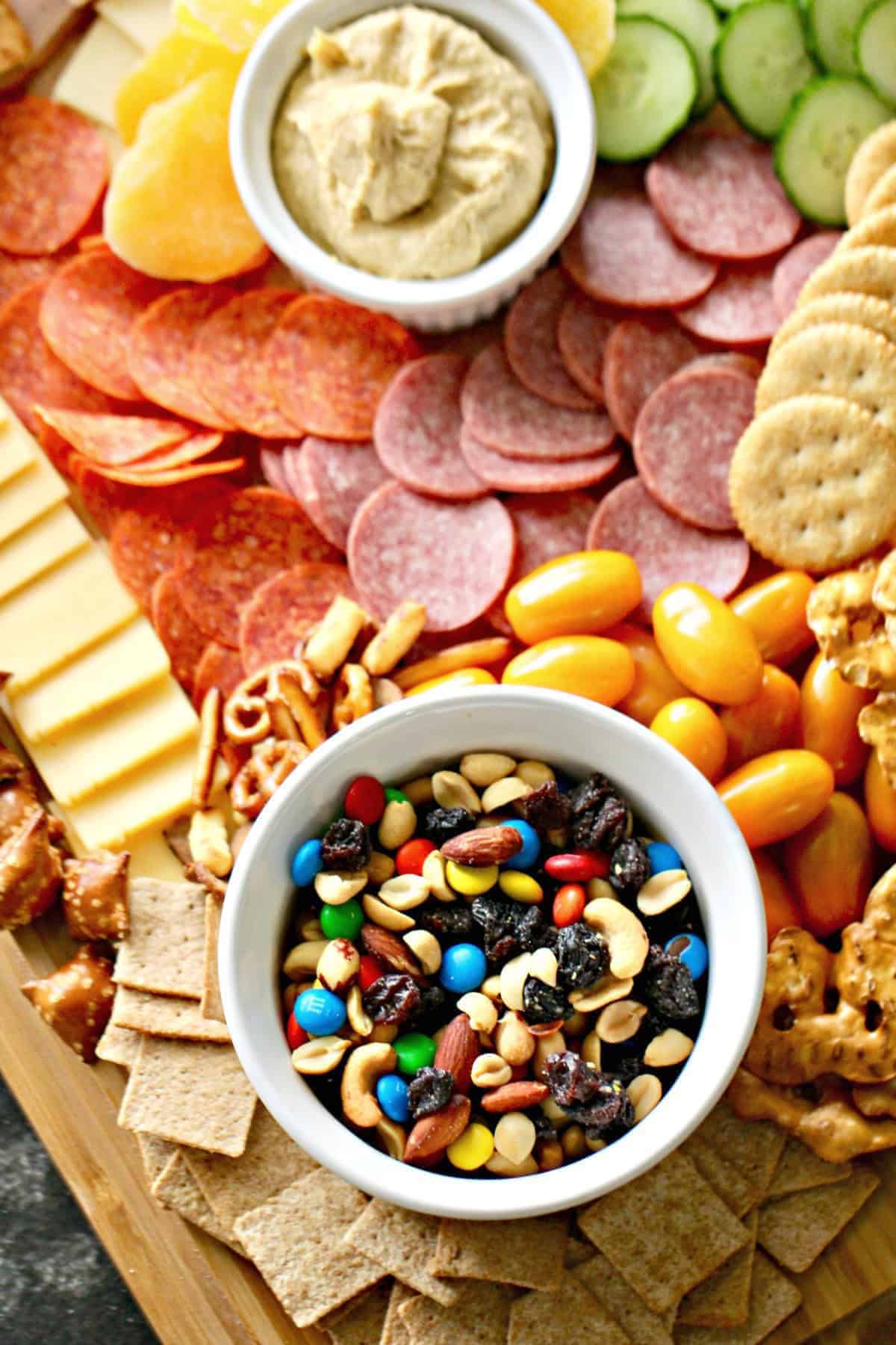 Charcuterie board with sliced meats, cheeses, crackers, pretzels, grape tomatoes, cucumber slices, dried fruits, nuts, and two bowls of dip and trail mix.