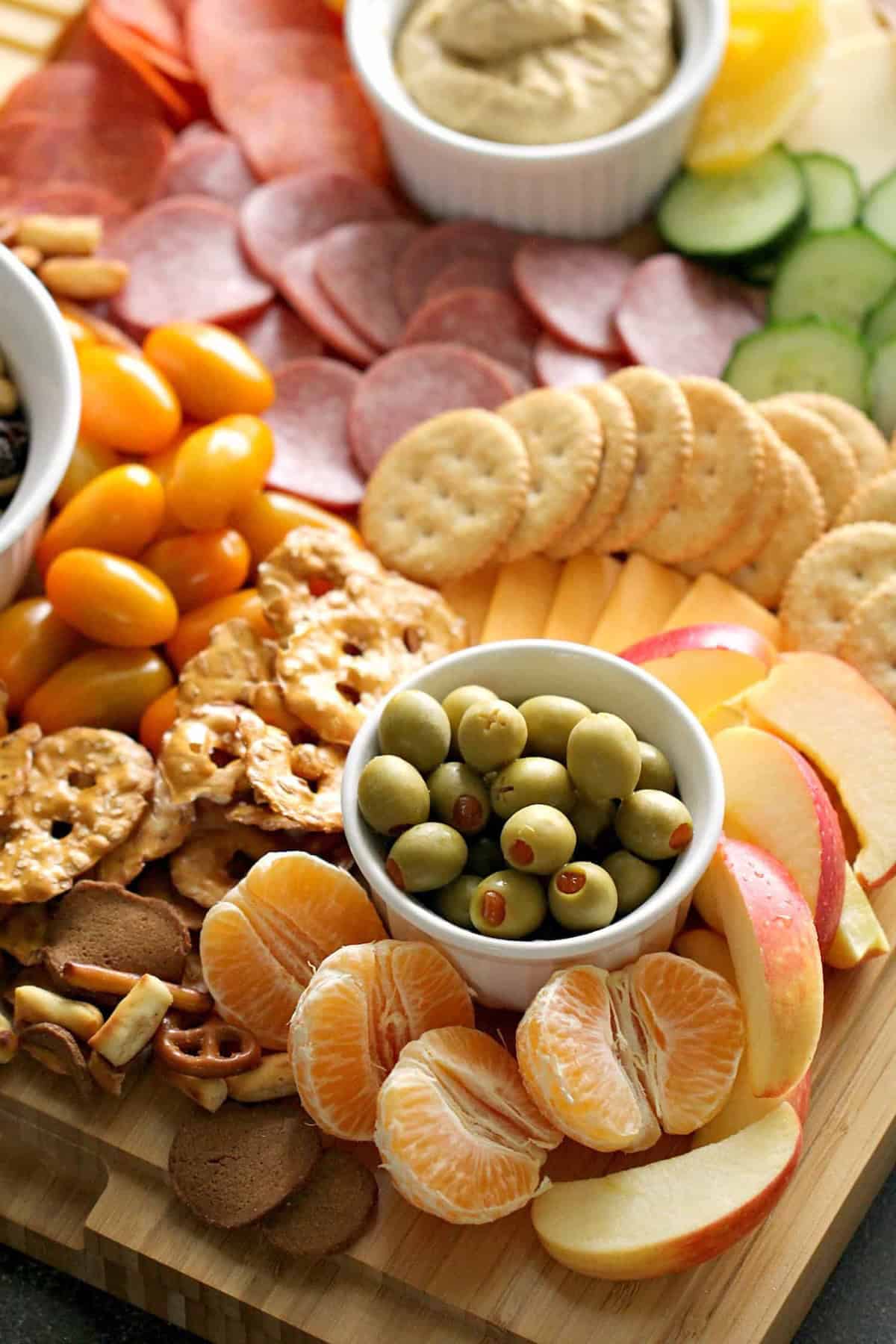 A charcuterie board with sliced meats, crackers, cucumber, cheese, olives, cherry tomatoes, pretzels, apple slices, tangerine wedges, and a dip.