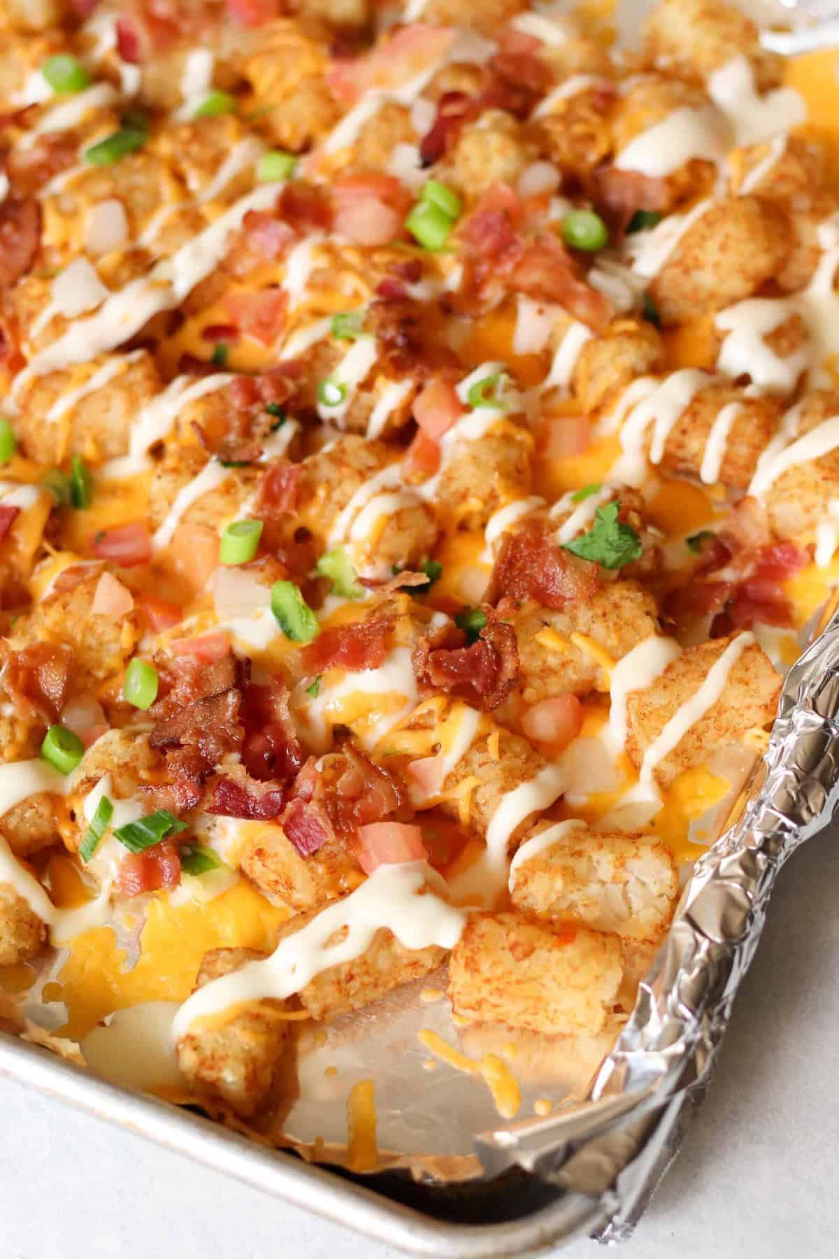 Image of loaded tater tots on a baking sheet, topped with melted cheese, bacon bits, chopped green onions, tomatoes, and drizzled with creamy sauce.