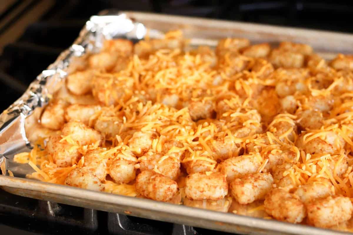 Baking sheet with tater tots covered in melted cheese, placed on a stove.