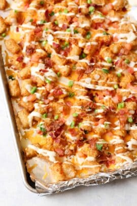 Tray of loaded tater tots topped with melted cheese, bacon bits, diced tomatoes, green onions, and drizzled with a white sauce.