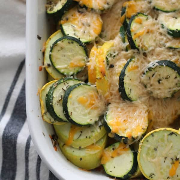 A baked dish of sliced zucchini and yellow squash topped with melted cheese in a white casserole dish, placed on a striped cloth.