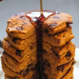 A stack of thick pancakes with visible chocolate chips being drizzled with syrup.