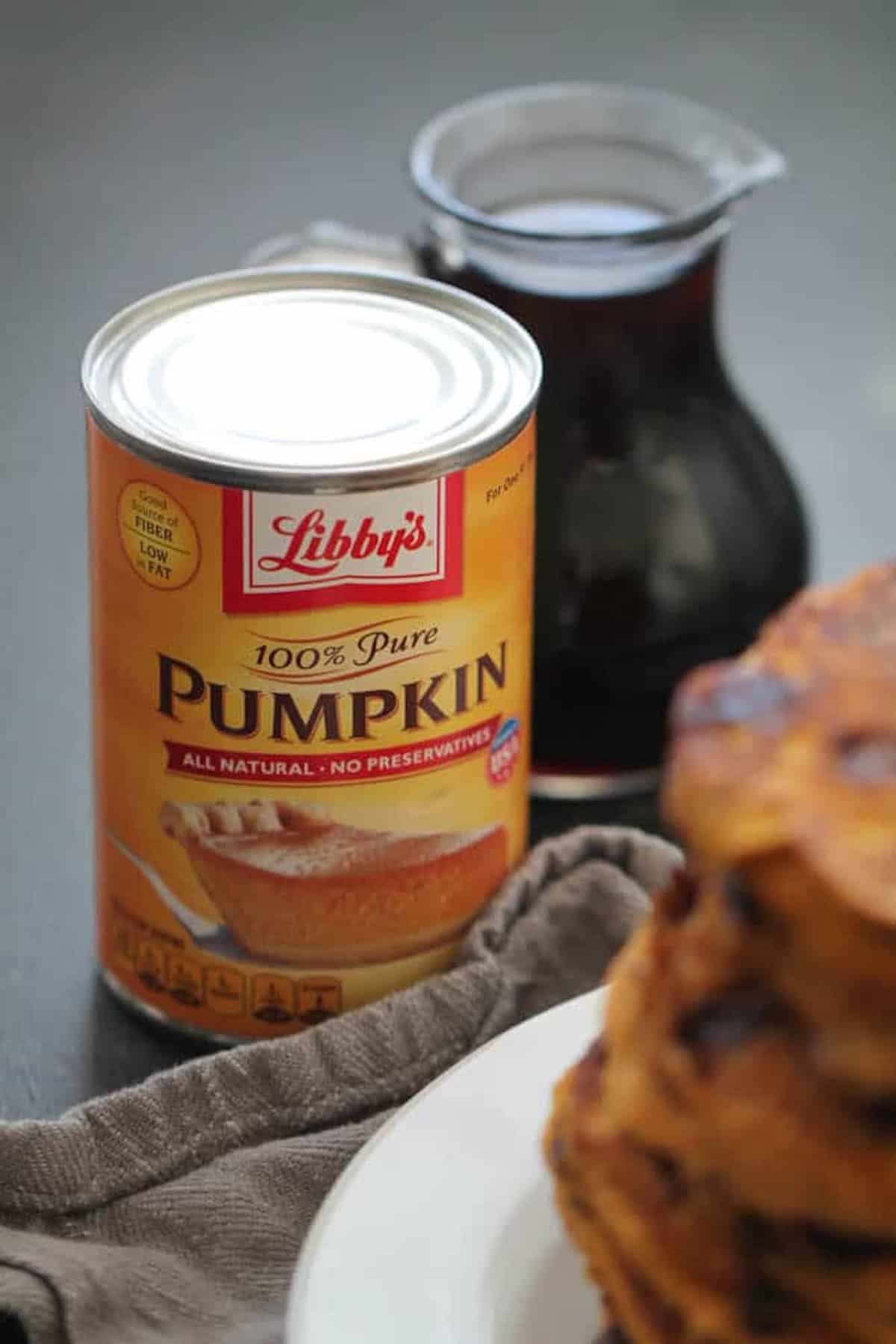 A can of Libby's 100% Pure Pumpkin with a stack of pancakes and a jug of syrup on a dark surface.