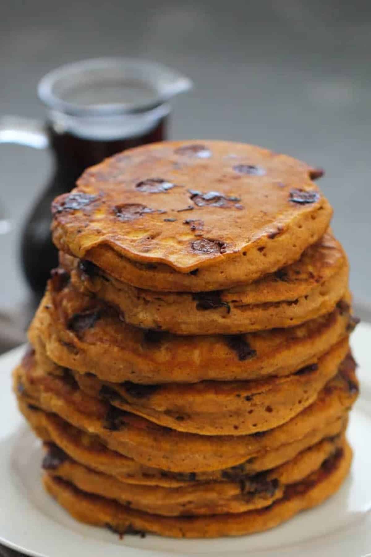 A stack of seven thick, golden-brown pancakes with visible chocolate chips, placed on a white plate. A glass pitcher filled with dark syrup is in the background.