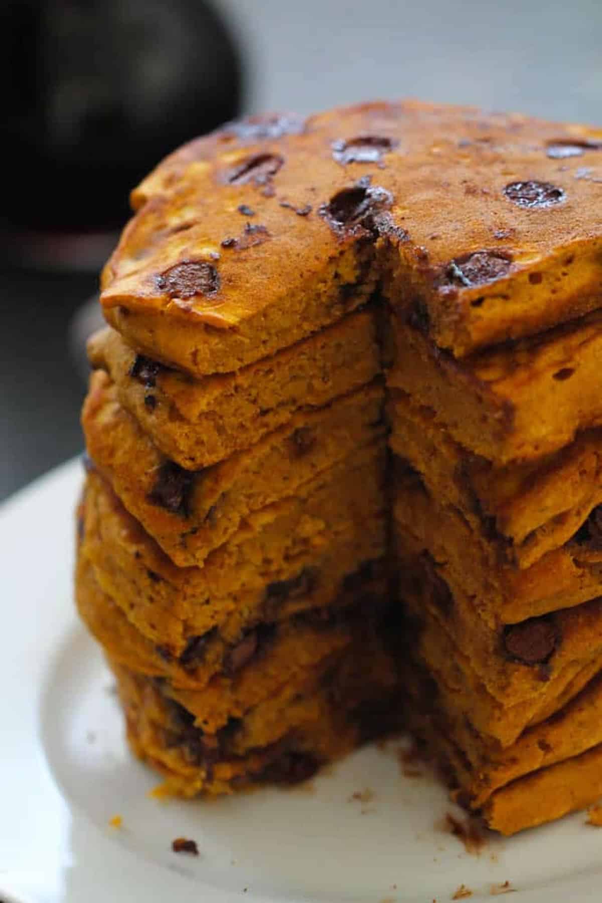 A tall stack of pumpkin chocolate chip pancakes with a slice cut out, revealing the dense texture and chocolate chips inside.