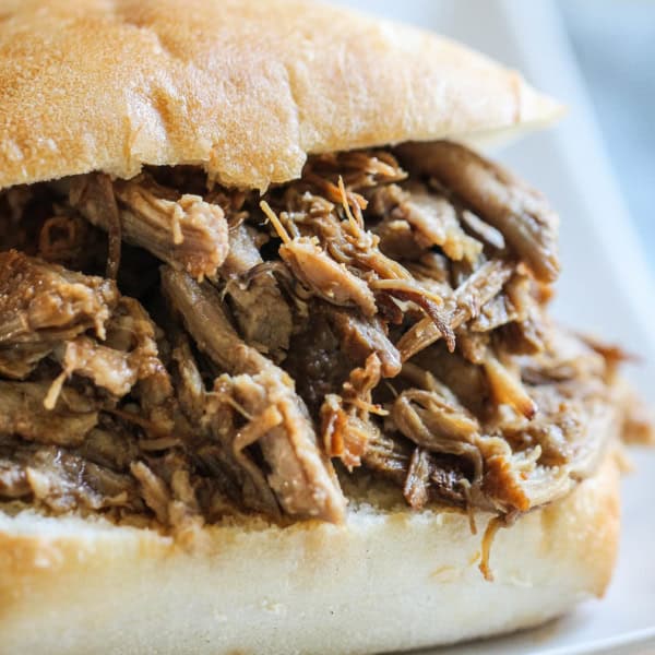 A close-up of a pulled pork sandwich served on a white plate. The sandwich has shredded pork meat inside a bun.