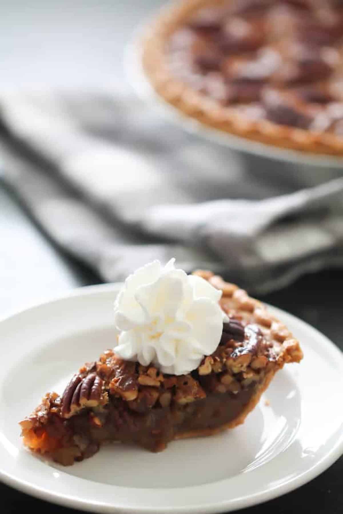 A slice of pecan pie topped with whipped cream on a white plate, with a whole pie in the background.