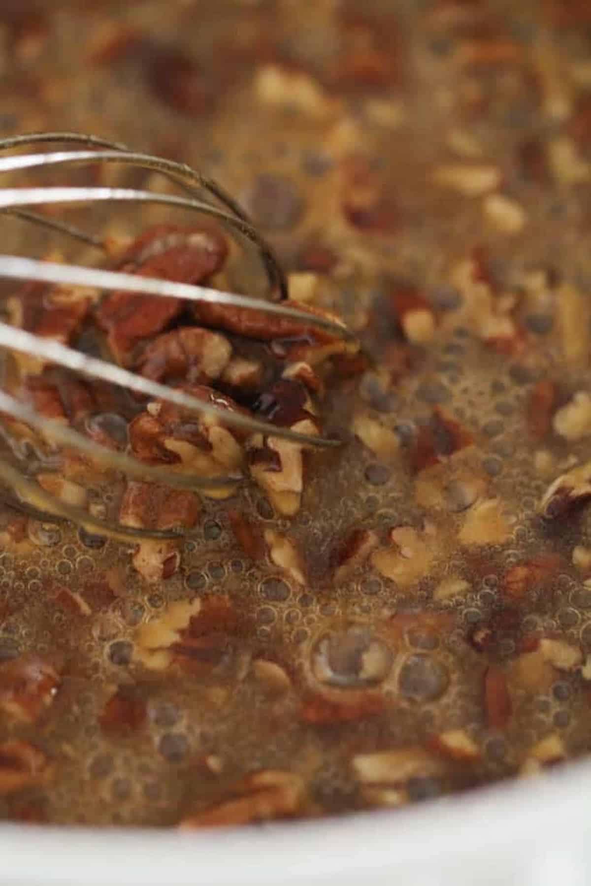 Close-up of chopped pecans being mixed with a whisk in a brown liquid batter.