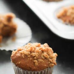 A freshly baked muffin with a crumbly topping sits in a white paper liner. More similarly topped muffins are visible in a baking tray in the background.