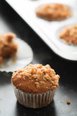 A freshly baked muffin with a crumbly topping sits in a white paper liner. More similarly topped muffins are visible in a baking tray in the background.