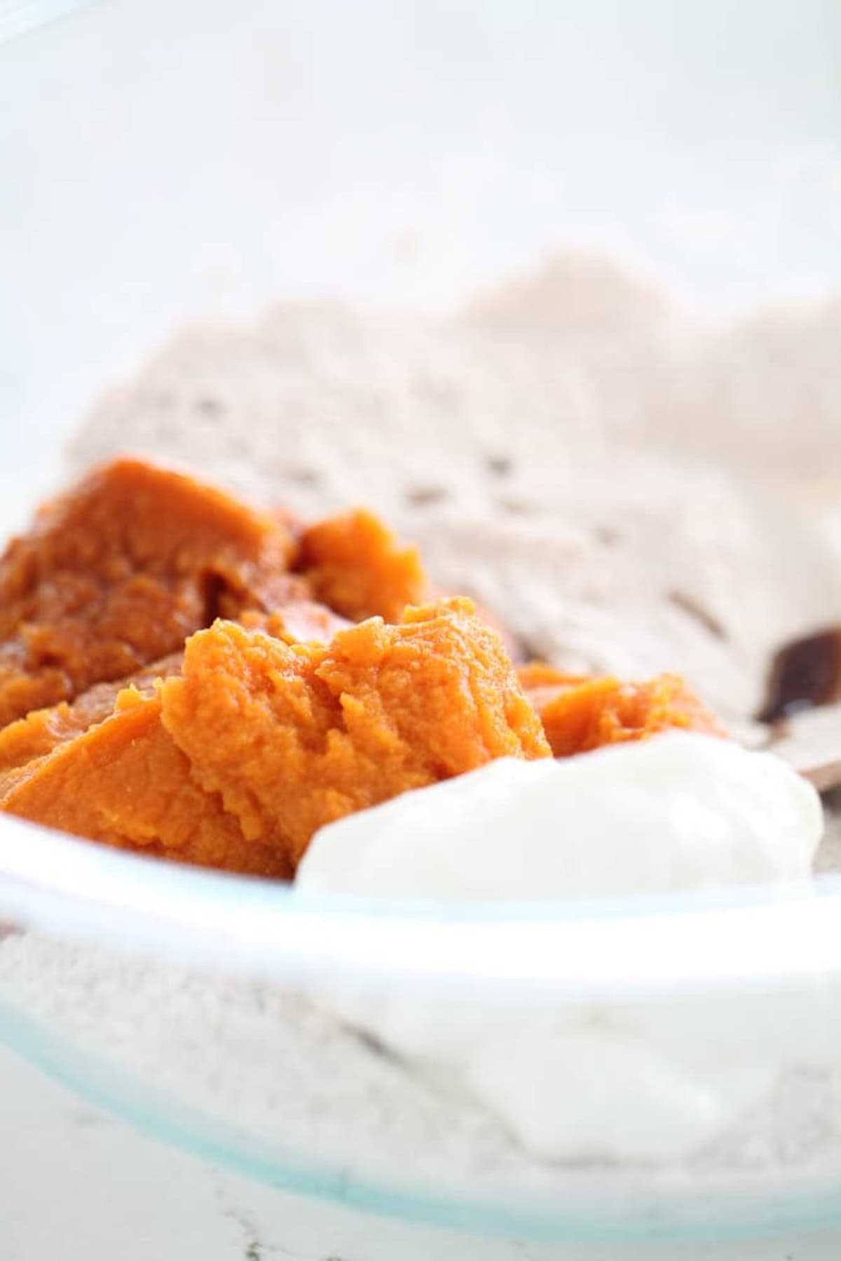 A glass mixing bowl containing pumpkin puree, yogurt, and a mixture of dry ingredients for baking.