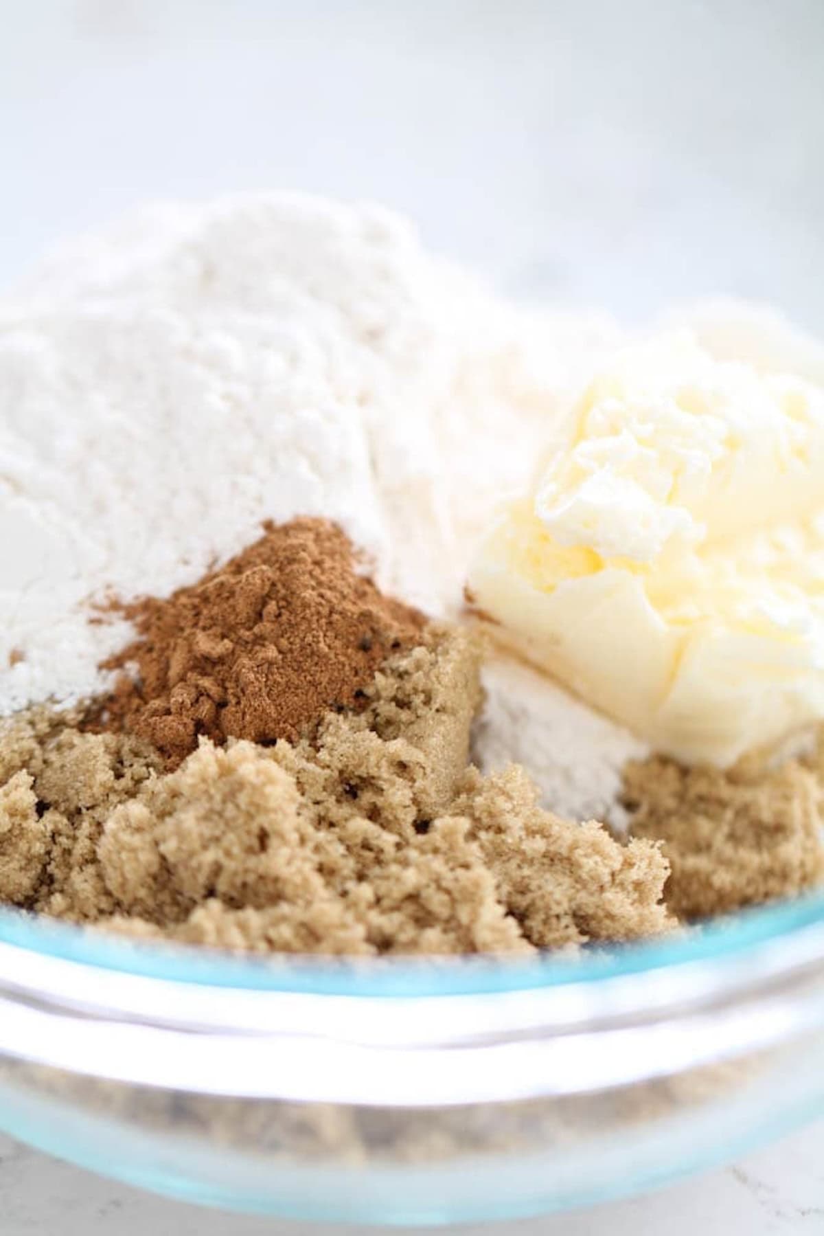 Close-up of a glass bowl with flour, brown sugar, a sprinkle of cinnamon, and a chunk of butter, all unmixed.