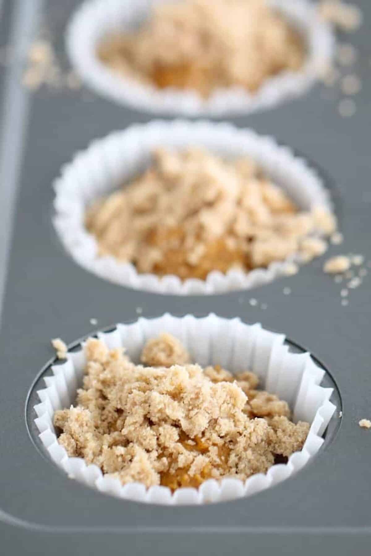 Unbaked muffins in white paper liners placed in a gray muffin tin, topped with a crumbly streusel mixture.