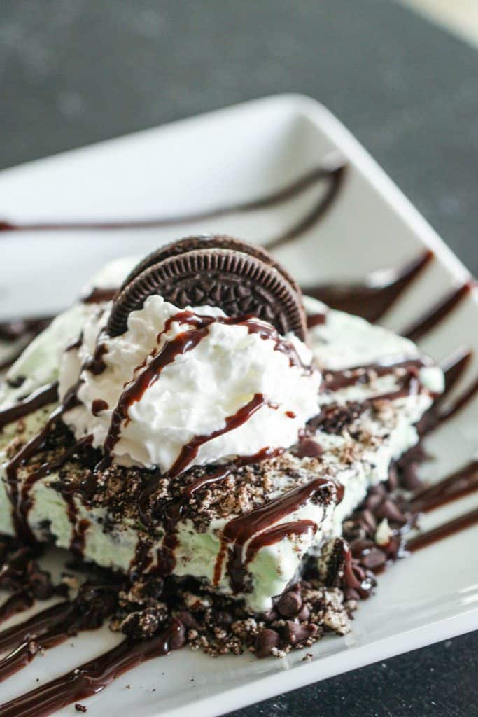 A slice of Asphalt Sheet Cake Pie on a white plate drizzled with chocolate sauce and topped with whip cream and an oreo cookie.