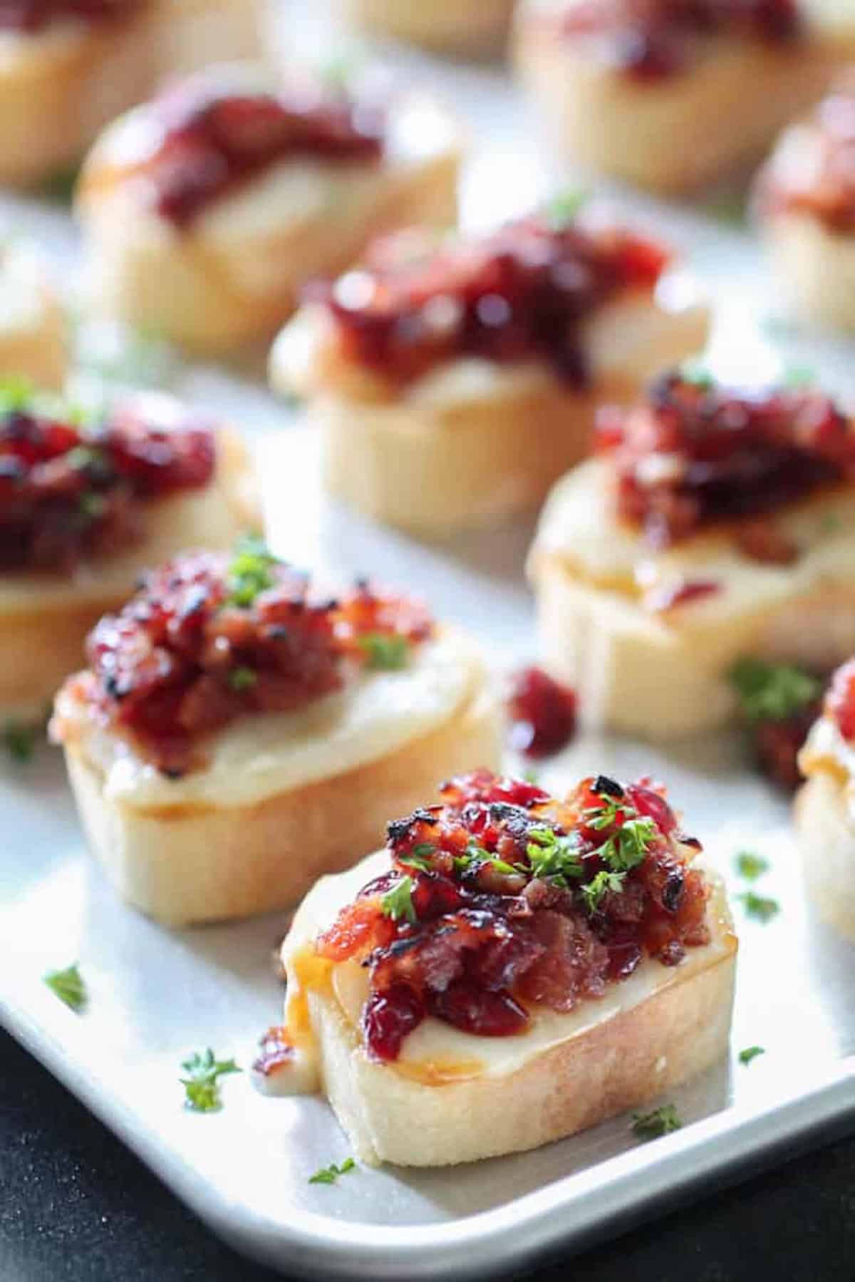 A close-up of several small bread slices topped with melted cheese, a jam or relish, and garnished with chopped herbs, arranged on a white surface.