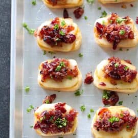 Tray of baked cranberry bacon jam crostinis topped with cheese, bacon, and parsley.