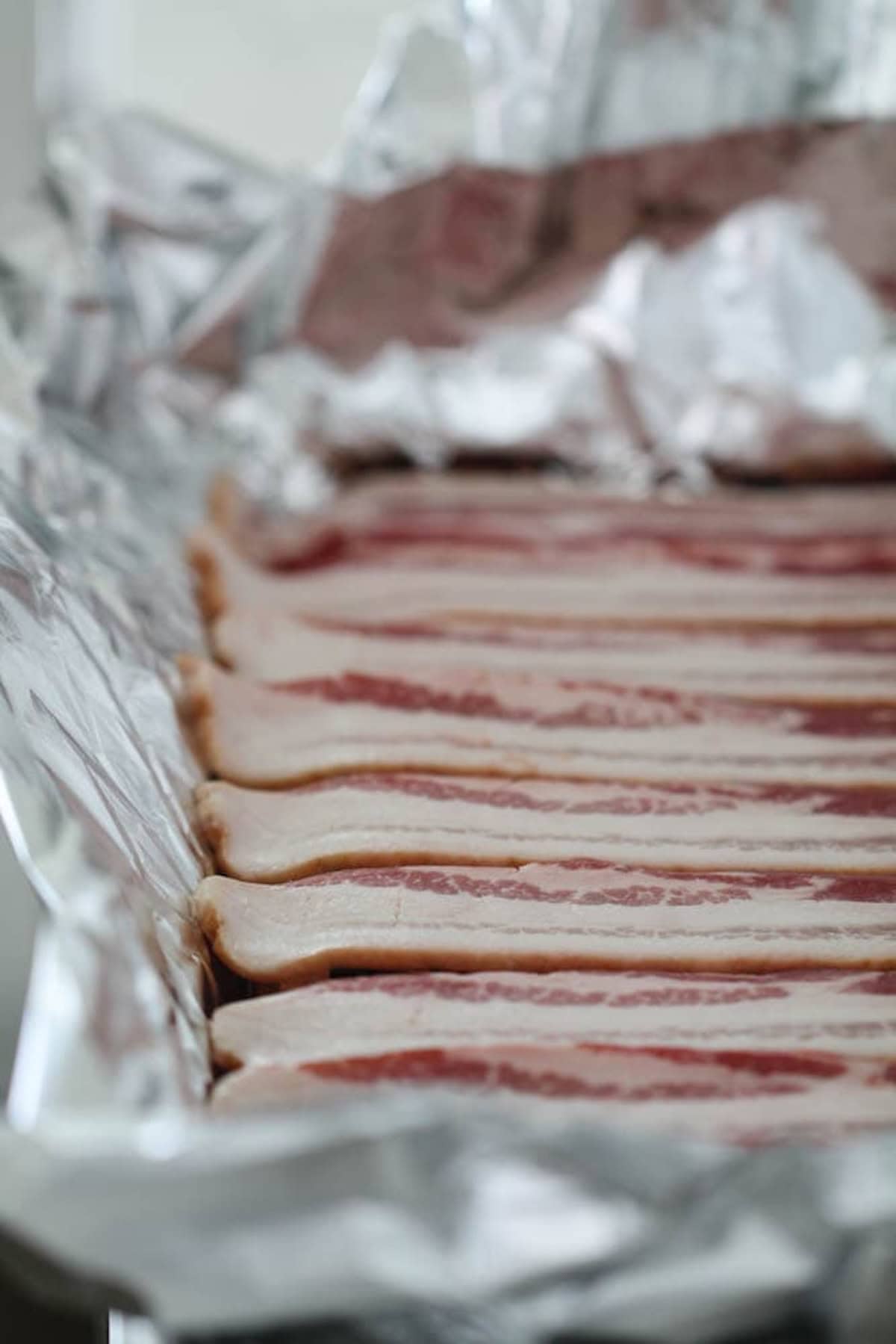 Strips of uncooked bacon are arranged neatly on aluminum foil.