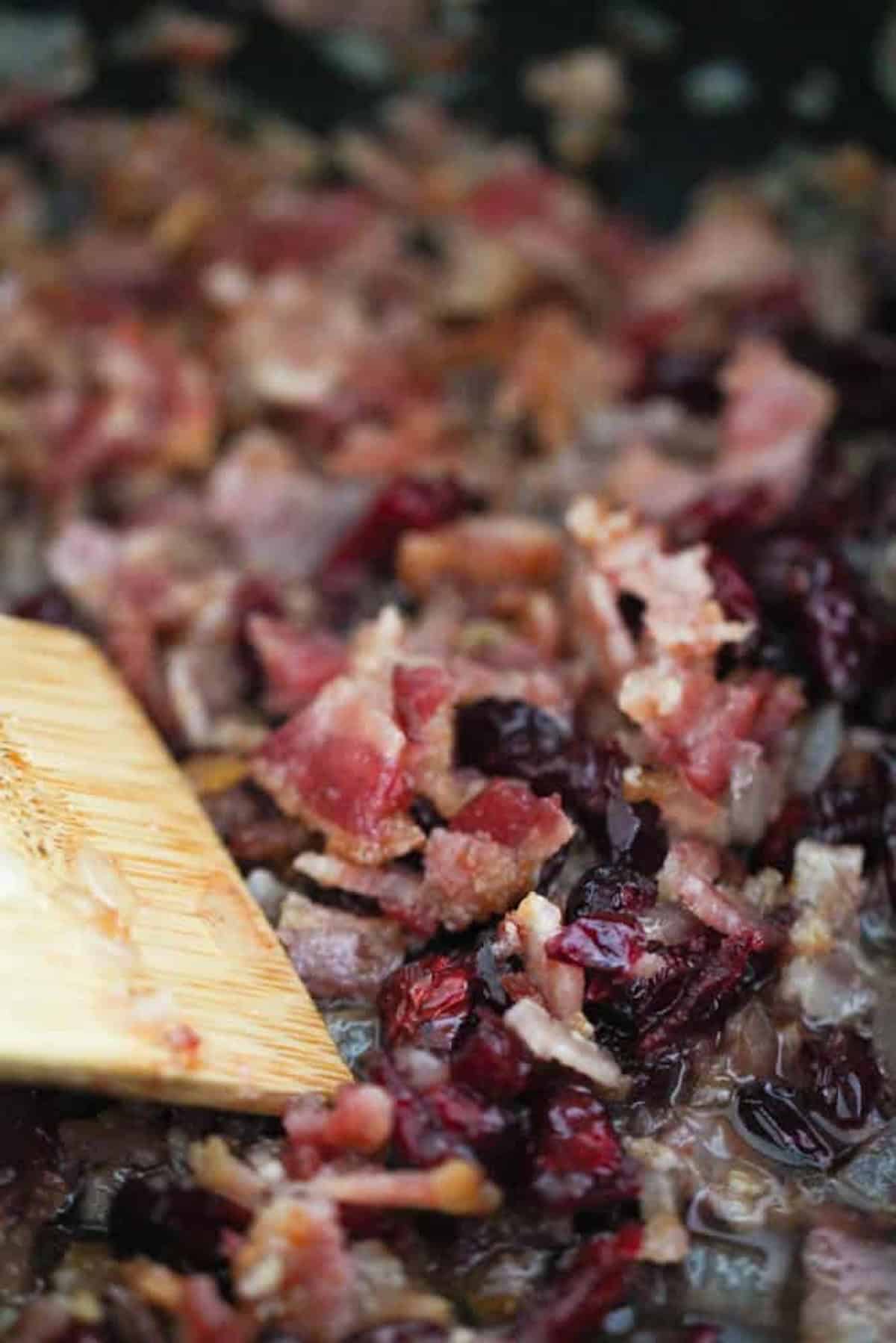 Chopped bacon bits and cranberries being stirred in a pan with a wooden spatula.