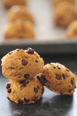 Three chocolate chip cookies stacked on a flat surface, with more cookies blurred in the background.