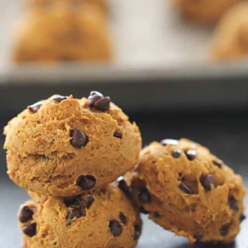 Three chocolate chip cookies stacked on a flat surface, with more cookies blurred in the background.