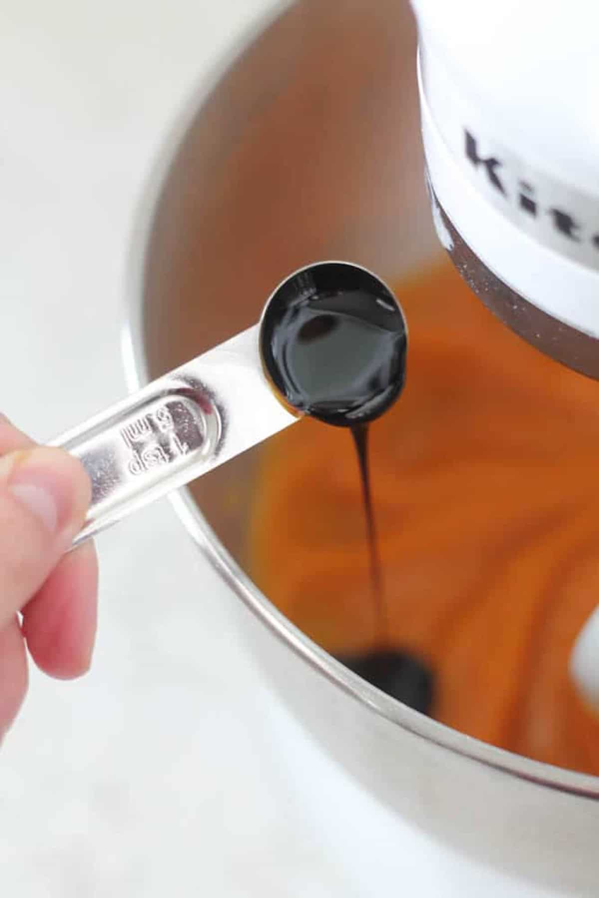 A close-up of a person adding liquid from a teaspoon into a mixing bowl attached to a stand mixer.