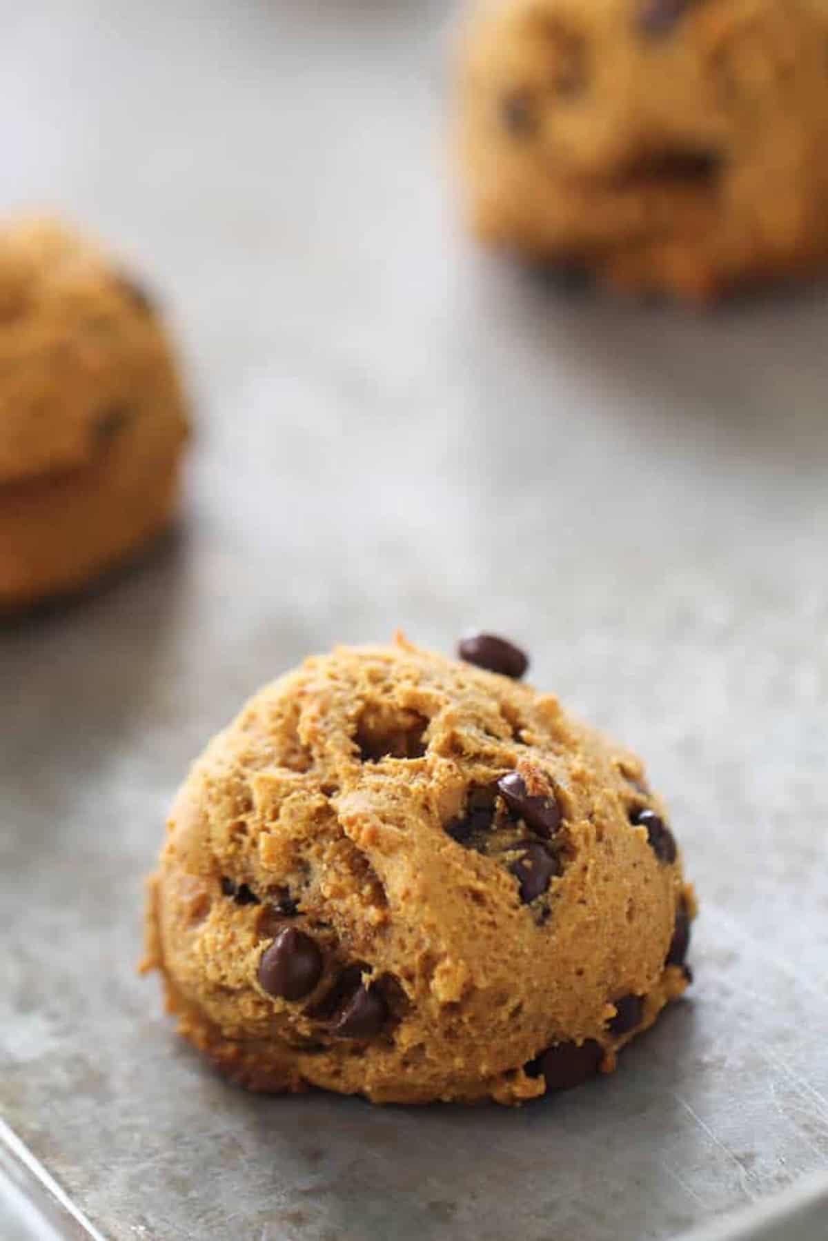 A close-up of a round pumpkin chocolate chip cookie on a baking tray, with a couple of blurred cookies in the background.