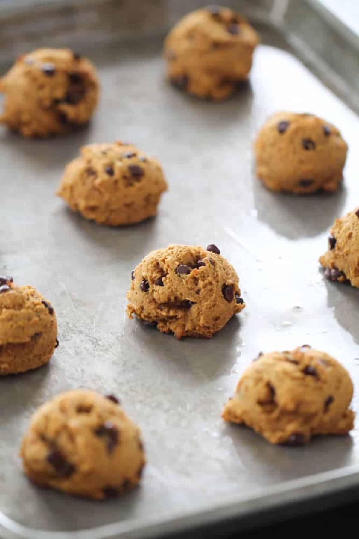 Nine dollops of chocolate chip cookie dough arranged in rows on a metal baking tray.