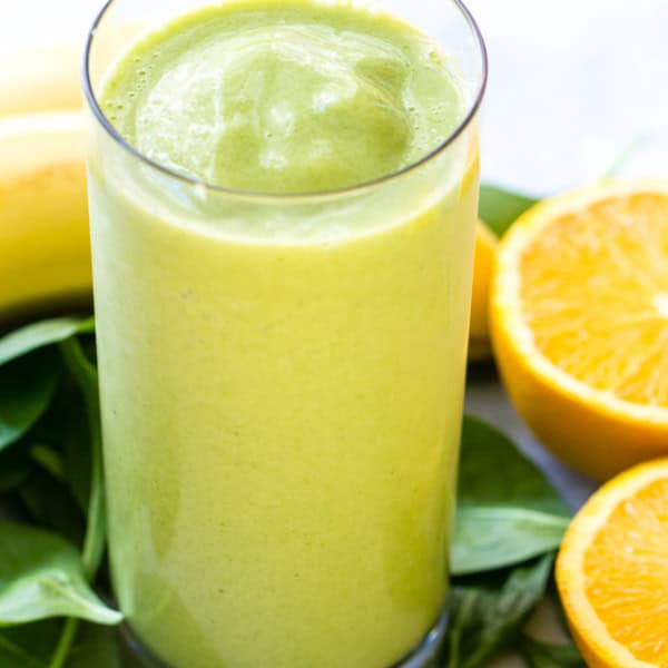 A tall glass of green smoothie is placed on a table with spinach leaves, a sliced orange, and a peeled banana in the background.