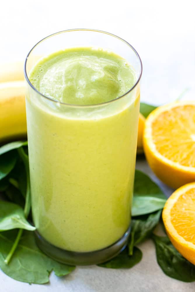 A tall glass of green smoothie is placed on a table with spinach leaves, a sliced orange, and a peeled banana in the background.