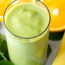 A glass of green smoothie is placed in the center, with orange slices and a banana in the background.