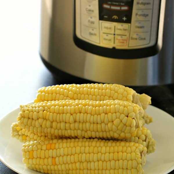 Four ears of corn on a white plate in front of an Instant Pot pressure cooker.