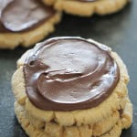 Three peanut butter cookies stacked, each topped with a layer of chocolate frosting, with more cookies in the background.