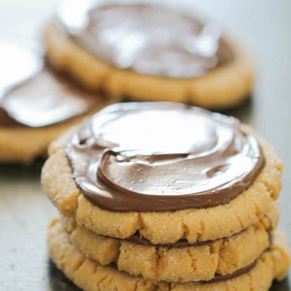 Three peanut butter cookies stacked, each topped with a layer of chocolate frosting, with more cookies in the background.