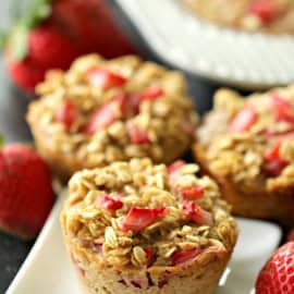 Three strawberry muffins with oat topping on a white plate, accompanied by fresh strawberries.