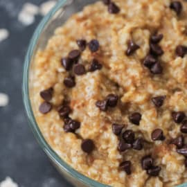 A glass bowl filled with oatmeal topped with chocolate chips. Scattered oats and a blurred can in the background.