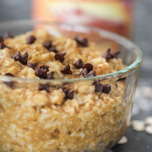 A glass bowl filled with oatmeal topped with chocolate chips. Scattered oats and a blurred can in the background.